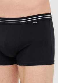 Primo piano di una persona che indossa boxer neri con una cintura a righe e un piccolo logo DIM sul lato sinistro.