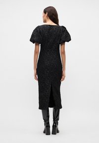 Črna obleka z napihnjenimi rokavi, cvetličnim teksturiranim materialom, do kolen dolga zasnova z razporkom na hrbtu, v kombinaciji z visokimi sijajnimi črnimi čevlji.