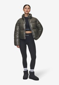 Dik olijfgroene jas met rechtopstaande kraag, zwart cropped top, zwarte leggings en zwarte gewatteerde schoenen. Witte sokken met logo.