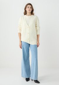 Cardigan crema a maglia cable con scollo a V, cinque bottoni e maniche lunghe, abbinato a jeans a gamba larga azzurri e scarpe nere a punta.