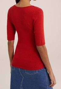 Rotes, geripptes Oberteil mit rundem Ausschnitt und 3/4-Ärmeln. Kombiniert mit einem Jeansrock, der eine figurnahe Silhouette und einen strukturierten Stoff zeigt.