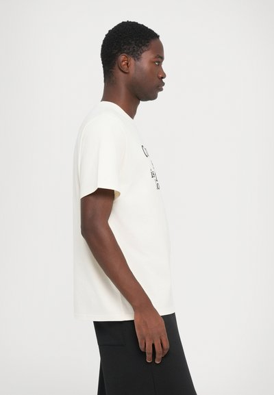 Crème-kleurige katoenen t-shirt met korte mouwen, met een bedrukt grafisch ontwerp op de voorkant. Het model draagt een zwarte broek en staat in profiel.