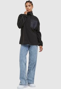 Pullover in pile nero con mezza cerniera, caratterizzato da una tasca nera. Abbinato a jeans a gamba larga azzurri e sneakers bianche.