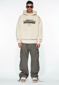 Felpa beige con tasca frontale e stampa grafica, abbinata a pantaloni cargo verdi con molte tasche e sneakers bianche con dettagli arancioni.