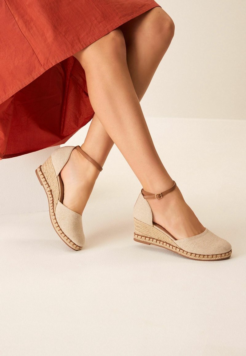 Jambes croisées portant des espadrilles compensées beiges avec des brides à la cheville et une jupe couleur rouille sur un fond clair uni.
