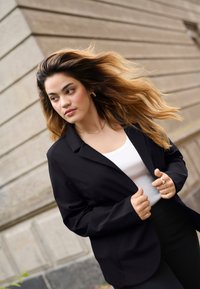 Blazer nero con vestibilità rilassata, indossato sopra una maglietta bianca. Tessuto morbido, dettagli minimi. Capelli lunghi e mossi che fluiscono nella brezza.