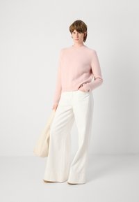Un pull en laine rose clair avec un col rond et des poignets côtelés, associé à un pantalon blanc à jambes larges. Un sac à main de teintes neutres posé sur le bras.