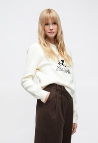 Balts džemperis ar apaļu apkakli un melnu grafisko apdruku. Kombinēts ar brūnām, augsta vidukļa biksēm. Mīksta faktūra, brīvs piegriezums.