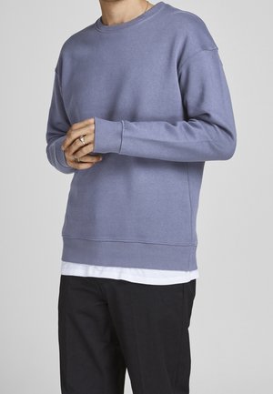 Sudadera de color púrpura claro, fabricada en un tejido suave, con cuello redondo, puños y dobladillo acanalados, superpuesta a una camisa blanca, combinada con un pantalón negro ajustado.