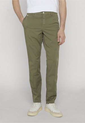 Homme portant un pantalon slim vert olive, des baskets blanches et une chemise blanche, debout avec une main dans la poche devant un arrière-plan uni.