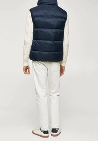 Tummansininen pehmeä liivi, jossa on korkea kaulus, pintakirjailtu valkoinen joustinneulepaita alla, yhdistettynä vaaleisiin farkkuihin ja rentoihin kenkiin.