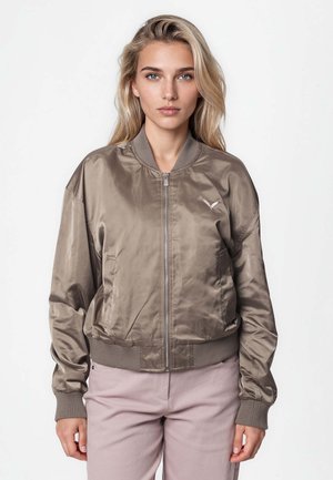 Lyshåret kvinde iført en taupe satin bomberjakke med ribbede manchetter og krave, stående mod en enkel hvid baggrund.