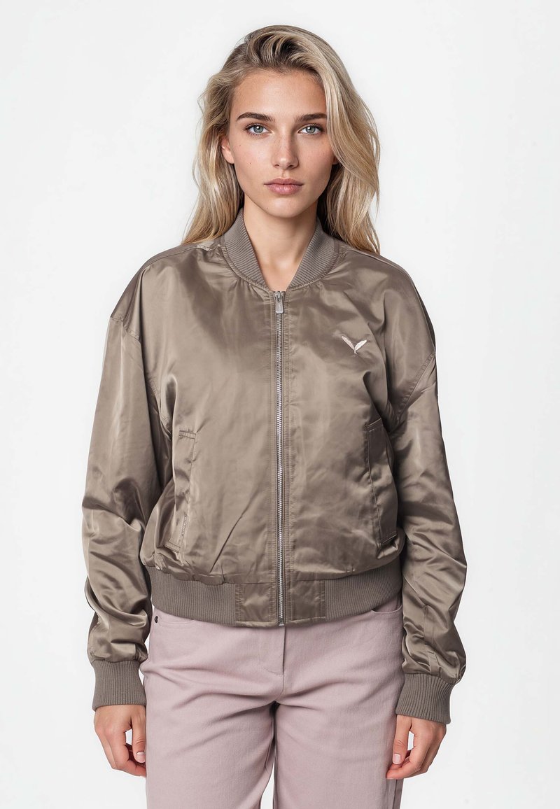 Femme blonde portant une veste bomber en satin taupe avec poignets et col côtelés, debout devant un fond blanc uni.