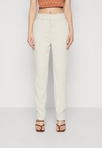 Pantalons beige ajustés avec une texture lisse, design à jambe droite, poches avant et taille mi-haute. Portés avec des sandales à lanières marron.