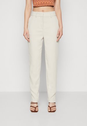 Pantalons beige ajustés avec une texture lisse, design à jambe droite, poches avant et taille mi-haute. Portés avec des sandales à lanières marron.