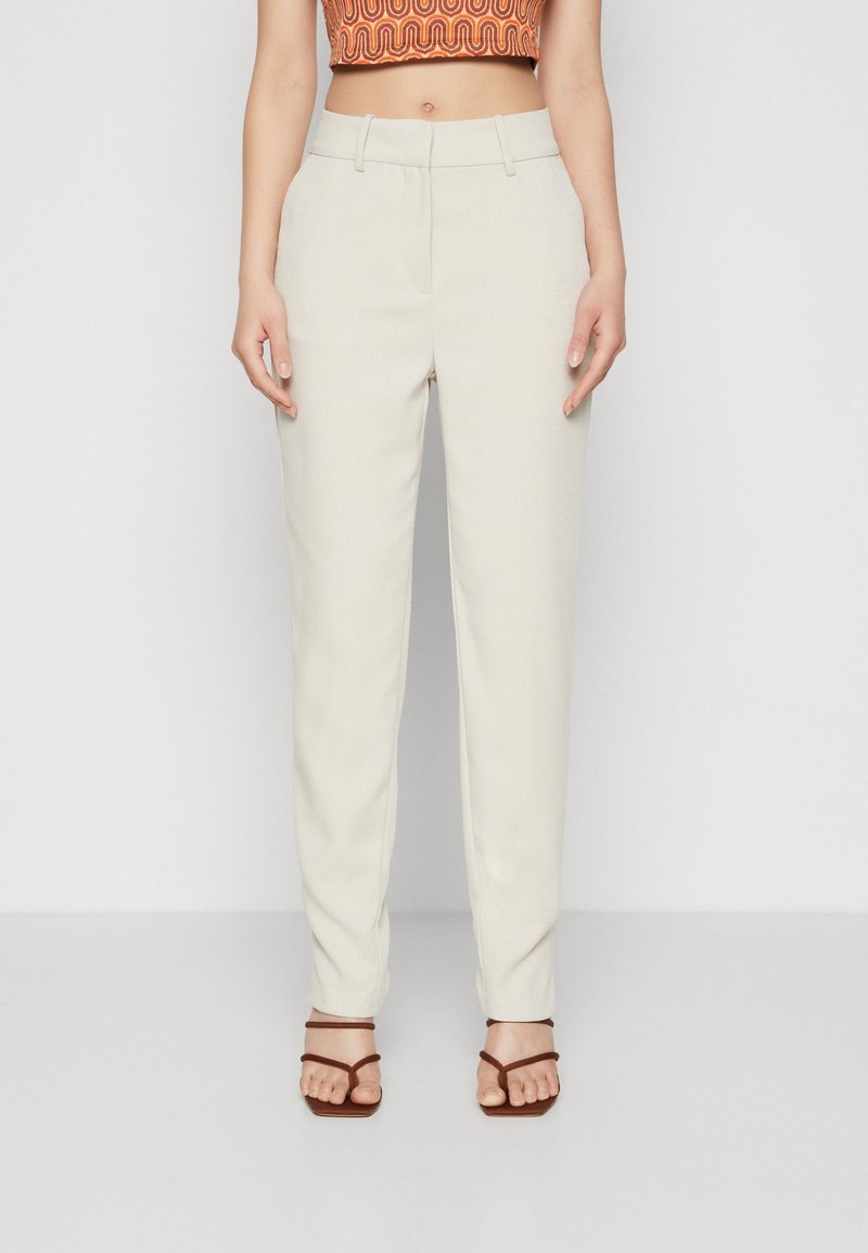 Pantalons beige ajustés avec une texture lisse, design à jambe droite, poches avant et taille mi-haute. Portés avec des sandales à lanières marron.