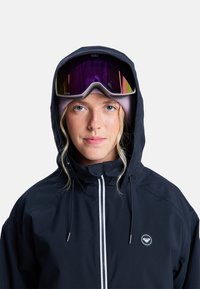 Young person wearing black hooded jacket, pink beanie, and ski goggles with reflective purple lenses, facing forward on plain background.