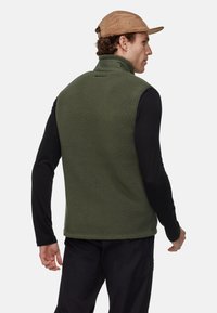 Groene fleece vest met een hoge kraag, gestructureerd oppervlak en een rechte zoom, gedragen over een zwart langemouwshirt. De hoed is van bruine corduroy.