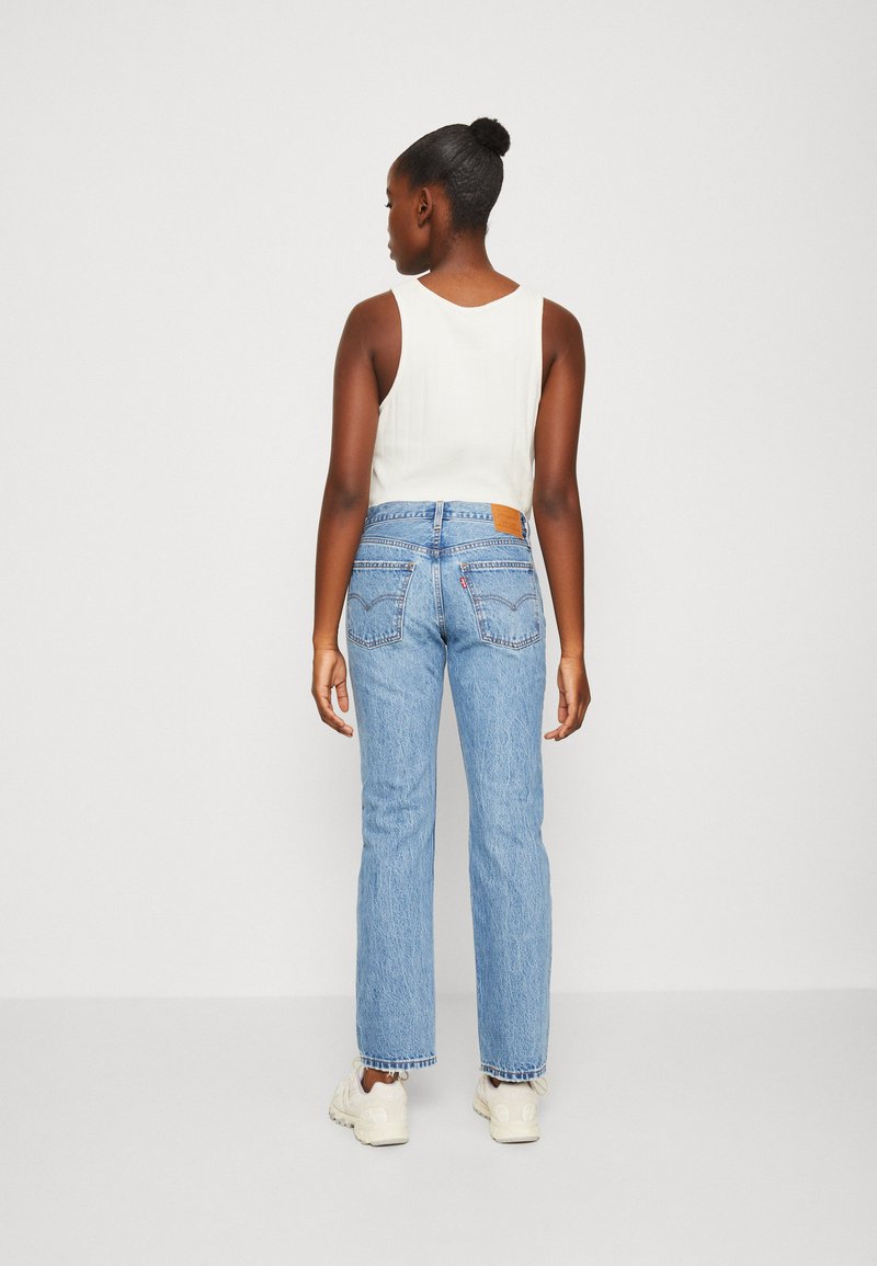 Leichtes, ärmelloses weißes Top kombiniert mit hochgeschnittenen, hellblauen Jeans aus Denim mit einem Lederaufnäher und geradem Bein.