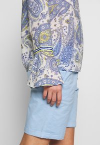Chemise à manches longues avec un motif paisley bleu et jaune, manches évasées et un poignet rayé. Associée à un short bleu clair.