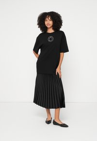 Mulher com cabelo encaracolado vestindo uma t-shirt preta oversized com um logotipo sorridente, saia midi preta plissada e sapatilhas pretas apontadas, com fundo branco.