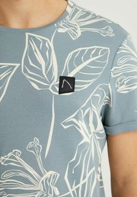 Gros plan d'un t-shirt bleu clair avec des contours floraux blancs et un logo carré noir sur la poitrine, porté par une personne à la peau claire.