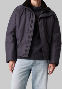 Mørkegrå isoleret jakke med kamskyderkrave, lynlås foran og trykknaplukninger, parret med en marineblå striktrøje og lyseblå jeans.