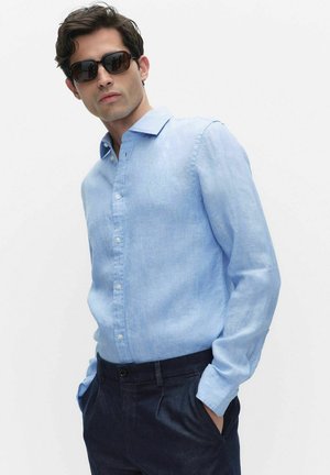 Mand iført mørke solbriller, lyseblå langærmet skjorte med knapper og mørke bukser, med hænderne i lommerne, står foran en ensfarvet baggrund.