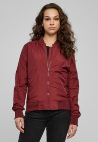 Veste bomber bordeaux avec fermeture éclair frontale, poignets et ourlet côtelés, finition en tissu lisse, poches latérales et détails froncés à la taille.