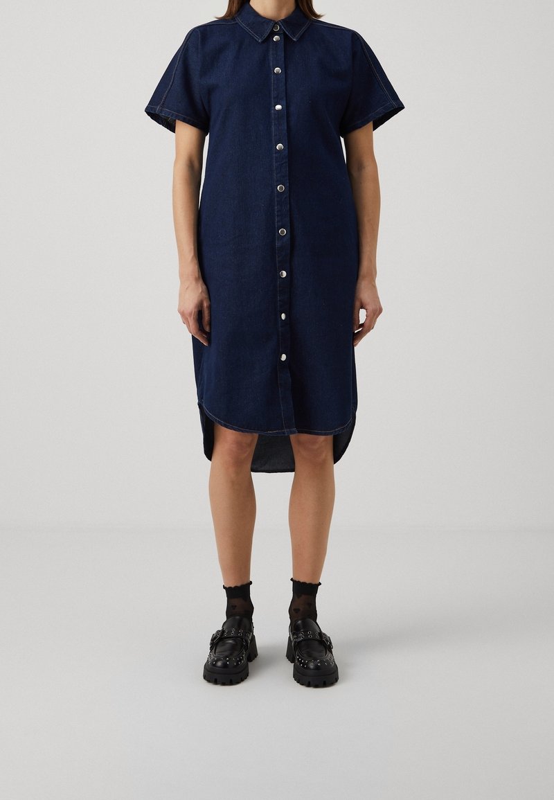 Mörkblå denimklänning med korta ärmar, knäppt framtill och böjd fåll. Bärs med svarta klumpiga skor och mönstrade strumpor.