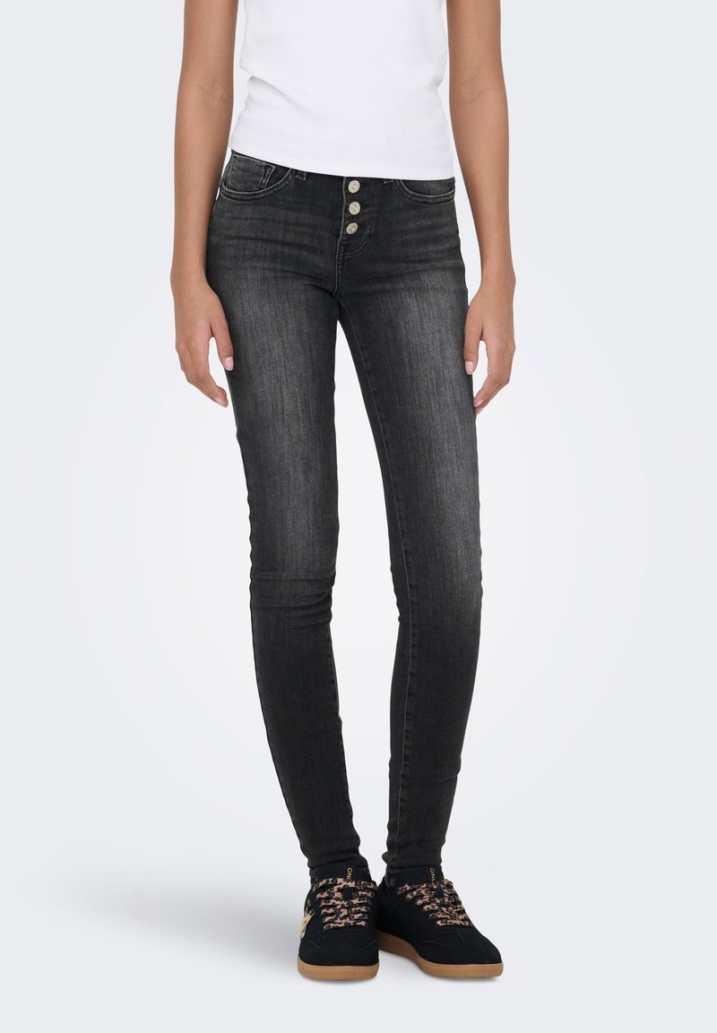 Άτομο που φορά ξεθωριασμένο στενό μαύρο τζιν με τέσσερα ασημένια κουμπιά και μαύρα αθλητικά παπούτσια με κορδόνια με σχέδιο σε λευκό φόντο.