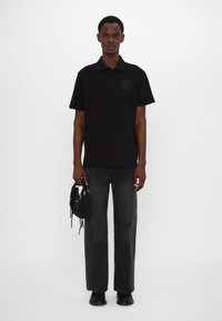Svart polo skjorte med kort krage, med en sirkulær logo på brystet. Bært med grå jeans og holder en liten svart veske.