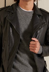 Veste de motard en cuir noir avec des éléments argentés, dotée d'une fermeture éclair asymétrique à l'avant et d'un pull gris en tricot en dessous.