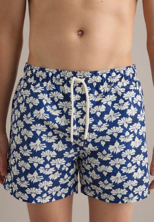 Pantaloncini da bagno blu con motivo floreale bianco e cordino bianco indossati da una persona con torso nudo su sfondo neutro.