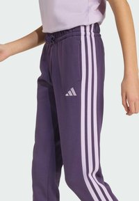 Fialové tepláky s elastickým pásom, bočnými vreckami a tromi bielymi pruhmi po stranách. Obsahujú malé biele logo na stehne.