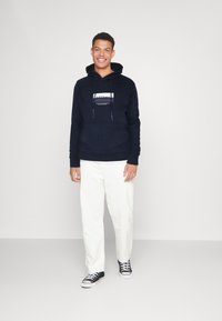 Felpa blu navy con tasca frontale, lacci e testo grafico. Abbinata a pantaloni bianchi e scarpe nere, in piedi di fronte a uno sfondo semplice.