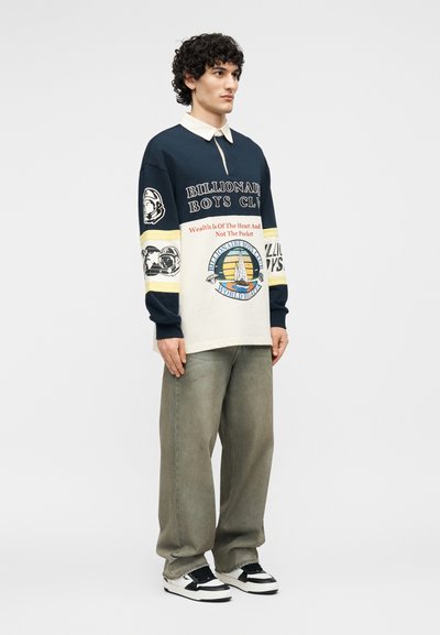 Νέος άνδρας με σγουρά μαλλιά στέκεται φορώντας μπλε σκούρο και λευκό μακρυμάνικο μπλουζάκι με λογότυπα και κείμενα, φαρδιά γκρίζα παντελόνια και ασπρόμαυρα αθλητικά παπούτσια.