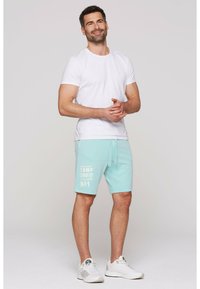 Witte crewneck t-shirt en licht turquoise shorts met een trekkoord en witte tekst. De shorts hebben het CAMP DAVID-merkteken. Witte sneakers.