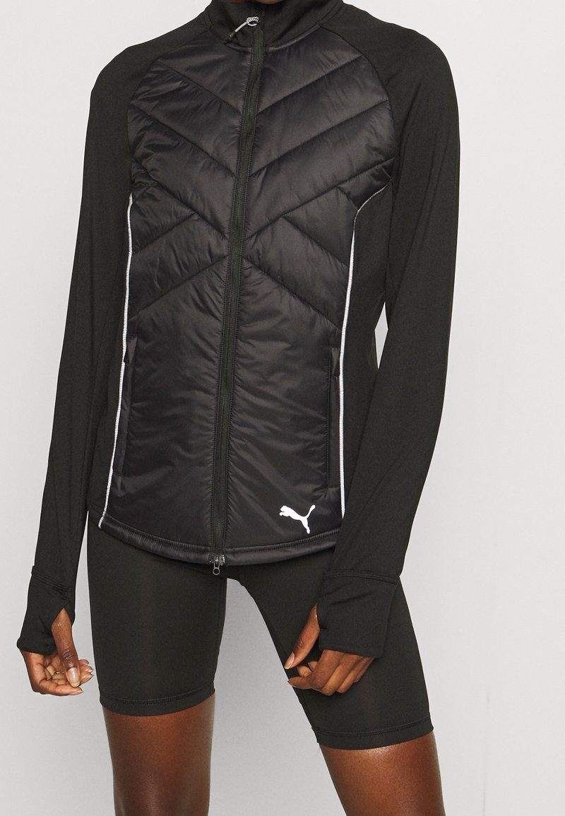 Gros plan d'une personne portant une veste matelassée noire zippée avec des trous pour les pouces et un short noir ajusté, montrant le logo Puma sur l'ourlet de la veste.