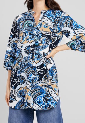 Femme portant une tunique à manches longues avec des motifs cachemire bleus, noirs, blancs et beiges et un jean bleu à jambes larges, posant avec une main sur la hanche.
