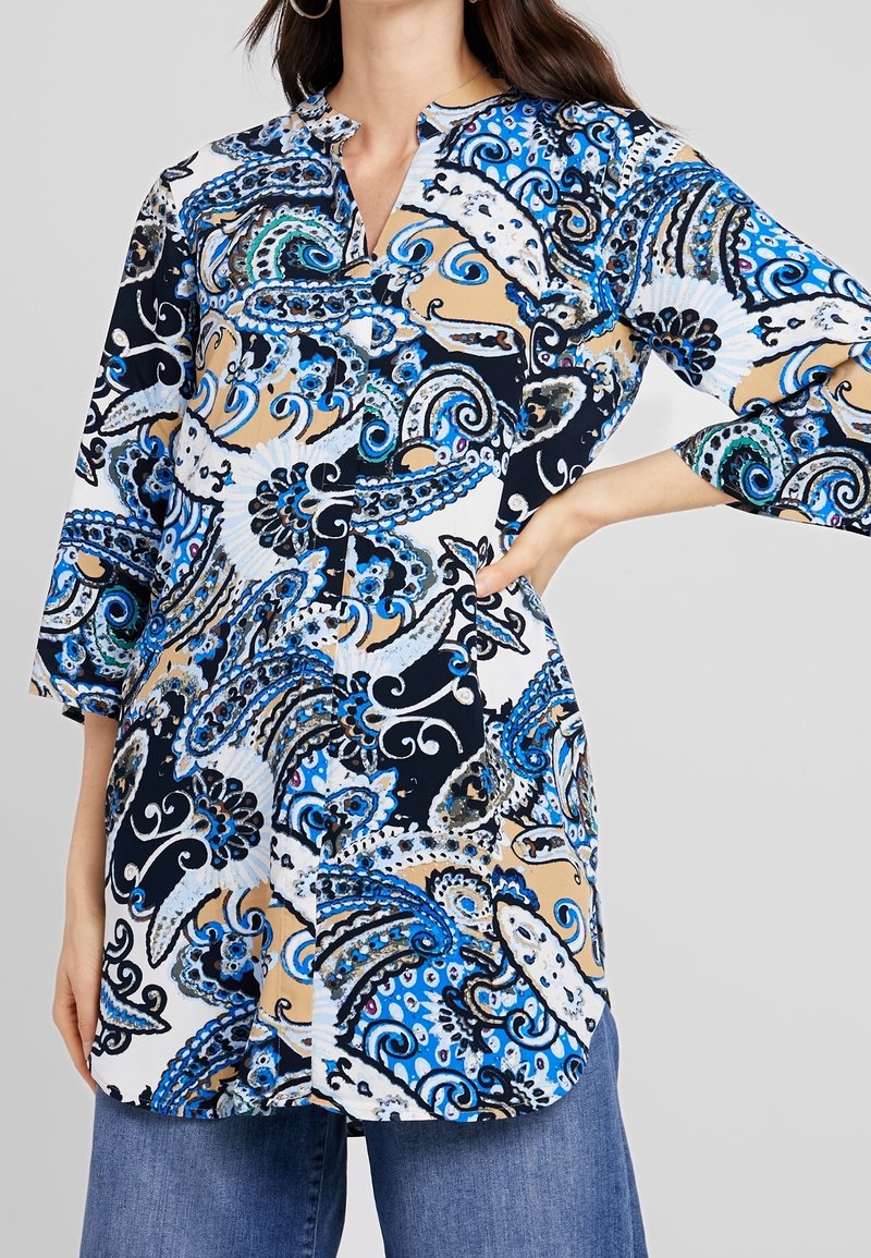 Femme portant une tunique à manches longues avec des motifs cachemire bleus, noirs, blancs et beiges et un jean bleu à jambes larges, posant avec une main sur la hanche.