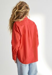 Person med langt bølget hår iført en løs rød skjorte med knapper og lyseblå jeans, vendt væk mod en ensfarvet lys baggrund.