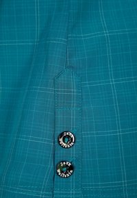 Tyrkysová košile na knoflíky s kostkovaným vzorem, svislou kapsou a dvěma kulatými  černými knoflíky s vyrytými detaily názvu značky.