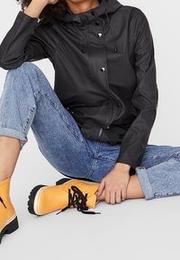 Czarna wodoodporna kurtka z kapturem, z zatrzaskami i sznurkiem do regulacji; w połączeniu z podwiniętymi niebieskimi dżinsami i jasnobrzoskwiniowymi botkami za kostkę.