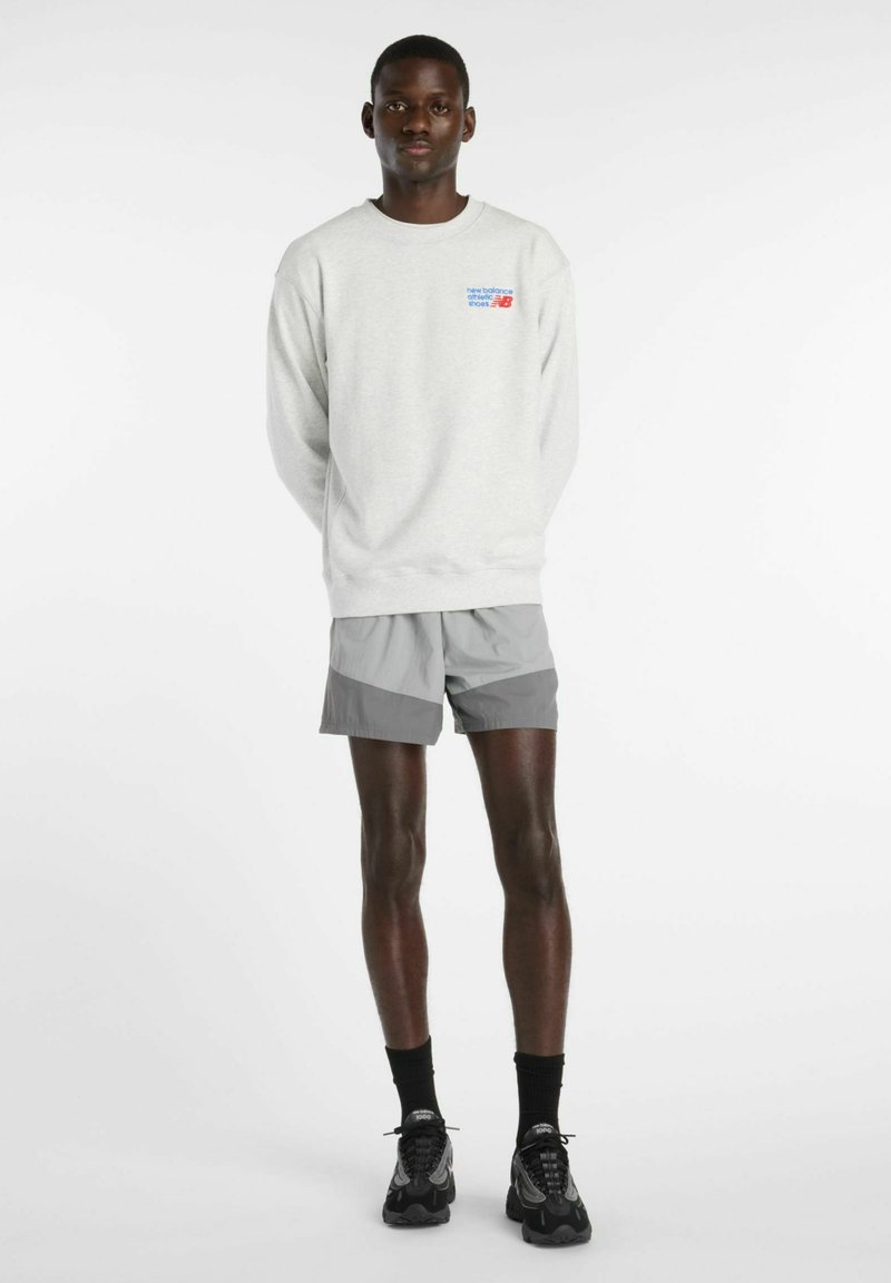 Mand stående med hænderne bag ryggen iført en lysegrå sweatshirt med logo, tofarvede grå shorts, sorte sokker og sorte sneakers.