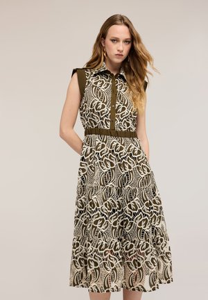 Kobieta o długich blond włosach, ubrana w sukienkę bez rękawów o długości do kolan z zielonym i białym kwiatowym wzorem koronkowym, stojąca z rękami w kieszeniach.