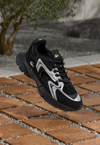 Zapatillas negras con acentos plateados, parte superior de malla, puntera redonda, suela texturizada y logotipos prominentes. Suspendidas sobre una superficie con patrón.