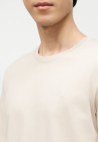 Κοντινό πλάνο προσώπου ατόμου που φοράει ανοιχτό μπεζ μπλουζάκι με στρογγυλή λαιμόκοψη και μικρό λευκό κεντημένο λογότυπο στο στήθος.