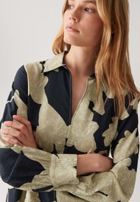 Junge Frau mit langen blonden Haaren, die eine beige und schwarze Bluse mit Blumendruck und langen Ärmeln trägt, mit verschränkten Armen zur Seite blickt.