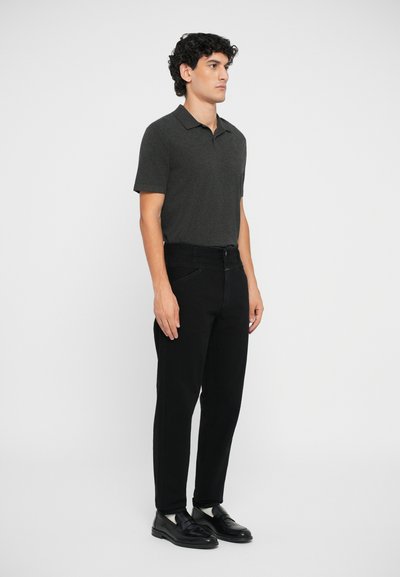 Cămașă polo gri cu mâneci scurte, asociată cu pantaloni negri slim-fit. Modelul stă drept, iar pantofii sunt negri cu un finisaj mat și fără modele vizibile.